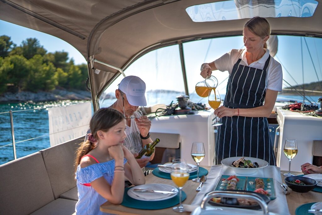 Die Gastgeberin schenkt einer Familie, die am Tisch auf dem Segelboot sitzt, Getränke ein